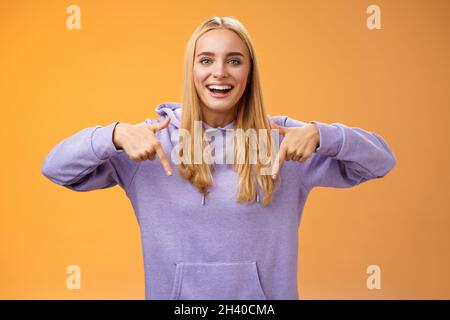 Amused joyful nice blond girlfriend pointing down present cool new product smiling broadly recommending try check out standing o Stock Photo