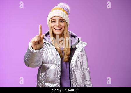 Excited cheerful fair-haired european girl in winter hat silver shiny jacket show number one index fingers give suggestion advic Stock Photo