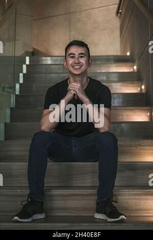 Asian young handsome man sitting on the stairs shop and smiling. Chinese guy, millennials life, lifestyle in the big city, Modern Asia, coffee break Stock Photo