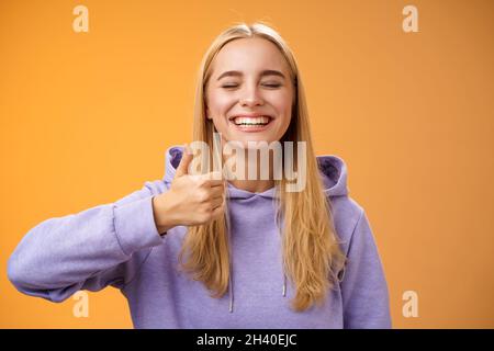 Amused joyful optimistic young excited blond european female model in hoodie smiling broadly close eyes having fun show approval Stock Photo