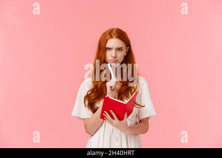 Serious-looking grumpy redhead woman frowning as thinking, pouting troubled, solving serious riddle, holding red notebook and to Stock Photo
