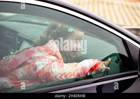 Little happy girl, child sitting inside the car seen from the outside. Young kid pointing outside the vehicle window, portrait, closeup. Means of tran Stock Photo