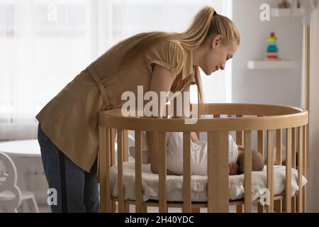 Loving young mother putting sleeping baby in wooden crib Stock Photo