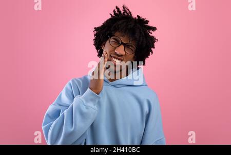Unhappy black teenager suffering from tooth ache, having gum problems, touching his cheek on pink studio background Stock Photo