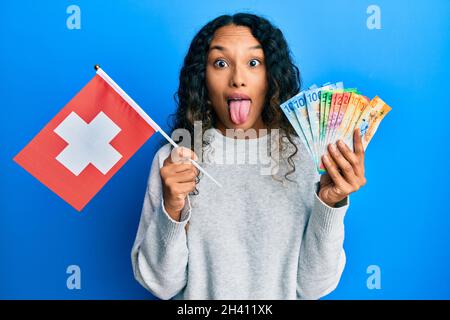 Young latin woman holding switzerland flag and swiss franc banknotes sticking tongue out happy with funny expression. Stock Photo