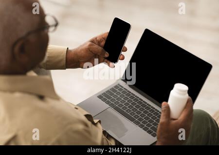 Senior African Man Using Phone And Laptop Holding Medication Indoors Stock Photo