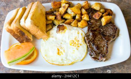 High protein breakfast plate: Sirloin stake and fried eggs. Sides dishes and garnish include potatoes, cantaloupe,  orange slice with toasts socked in Stock Photo