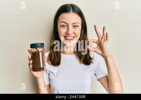 Young brunette woman holding soluble coffee doing ok sign with fingers, smiling friendly gesturing excellent symbol Stock Photo