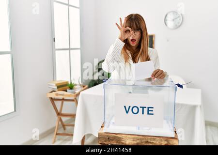 Hispanic business woman voting putting envelop in ballot box doing ok gesture shocked with surprised face, eye looking through fingers. unbelieving ex