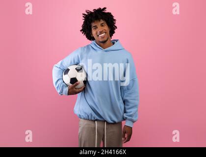 Portrait of positive black teenage guy holding soccer ball on pink studio background Stock Photo
