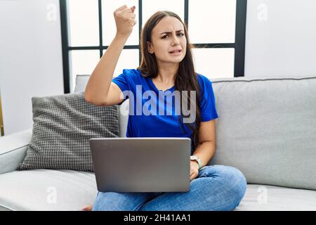 Young brunette woman using laptop at home angry and mad raising fist frustrated and furious while shouting with anger. rage and aggressive concept. Stock Photo