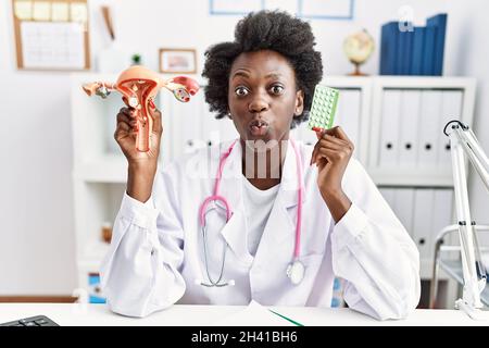 African doctor woman holding anatomical female genital organ and birth control pills puffing cheeks with funny face. mouth inflated with air, catching Stock Photo