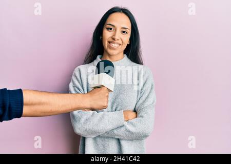 Young hispanic girl being interviewed by reporter holding microphone happy face smiling with crossed arms looking at the camera. positive person. Stock Photo