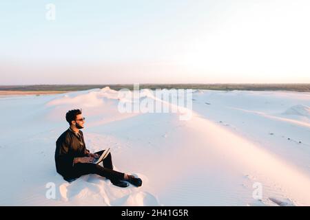 Handsome young Arab man writes on Internet using laptop and sits on sand amid sandy desert at sunset. Stock Photo
