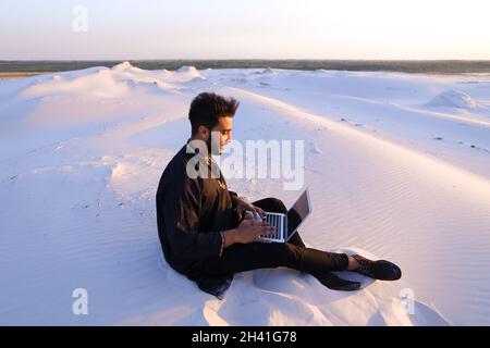 Handsome young Arab man writes on Internet using laptop and sits on sand amid sandy desert at sunset. Stock Photo