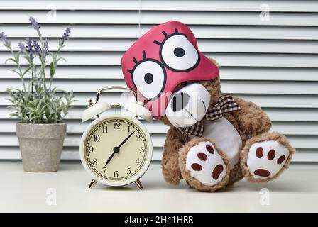 Brown teddy bear sits with a bandage for sleeping on a white table. Near the alarm clock, time is seven in the morning Stock Photo