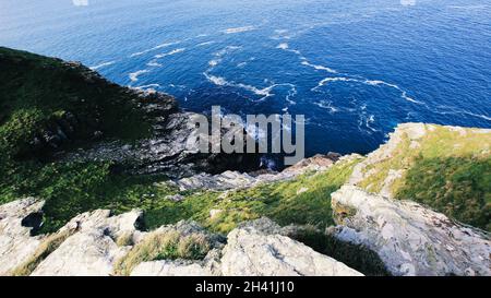 Cornish cliff side drop towards the sea, contrasting rock formations, plant life greens against the deep blue of open ocean. Stock Photo
