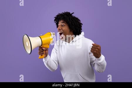 Emotional black adolescent shouting into megaphone, making announcement over violet studio background Stock Photo