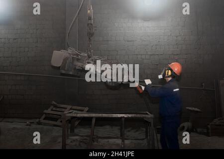 A working man in overalls, a protective helmet and a respirator controls heavy equipment for cleaning cast iron concrete tubing in the workshop of an Stock Photo