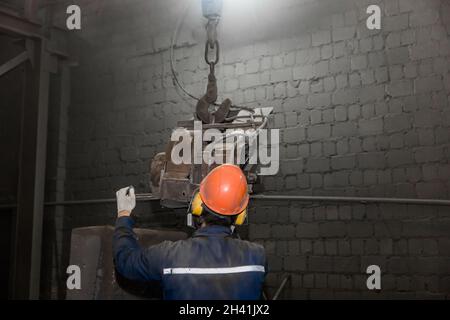 A working man in overalls, a protective helmet and a respirator controls heavy grinding equipment in the workshop of an industrial plant. Stock Photo