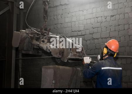 A working man in a protective helmet, respirator and overalls controls heavy grinding equipment for cleaning cast iron reinforced concrete structures Stock Photo