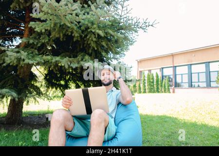 Prosperous man sitting in bag chair with laptop and looking at cam with close up face. Stock Photo