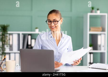 Busy female entrepreneur working on laptop and holding business reports, sitting at workplace in office interior Stock Photo