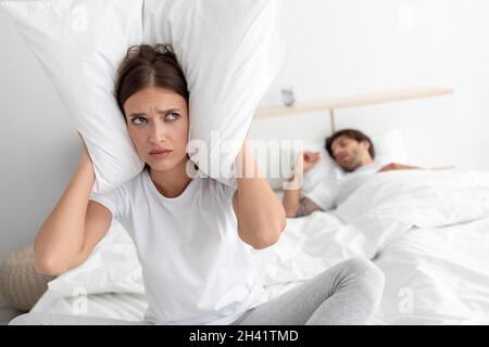 Tired unhappy caucasian wife covers ears with pillow sitting on bed with sleeping husband in bedroom interior Stock Photo