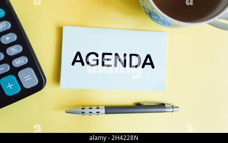 The word is AGENDA written on a notepad and a yellow background, next to a cup and a calculator Stock Photo