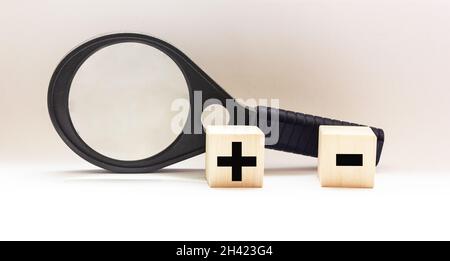 Two wooden cubes with mathematical symbols plus and minus, near a magnifying glass on a white background Stock Photo