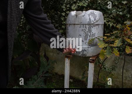 Man turning on the water tap on a white plastic water collection barrel at the green garden. Stock Photo