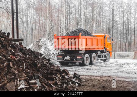 A dump truck in an industrial area or on a construction site in winter unloads coking coal from the body, next to a pile of metal waste and garbage. Stock Photo