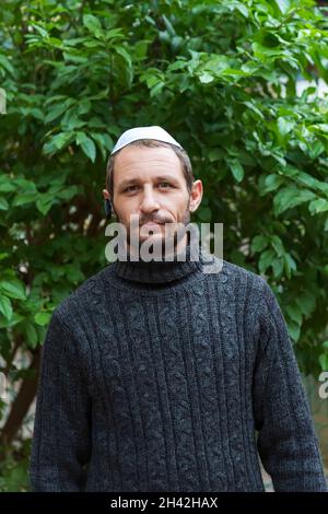 Jewish man wearing kippah in Hebrew or yarmulke (cloth cap traditionally worn by Jewish males) with green trees in the background, beard wearing black Stock Photo