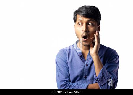 A young Indian male in blue dress with surprised shocked look and mouth opened looks sideways on white background Stock Photo