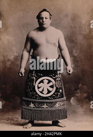 A young sumo wrestler posing in a photographic studio, wearing a richly decorated apron, in front of a painted backdrop. Photograph, ca.1900. Stock Photo