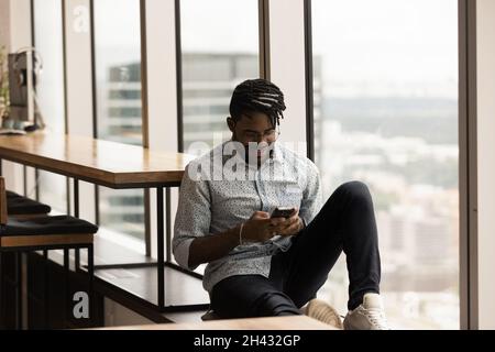 Relaxed african ethnicity man using smartphone, sitting on windowsill. Stock Photo