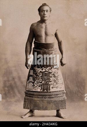 A young sumo wrestler posing in a photographic studio, wearing a richly decorated apron, in front of a plain backdrop. Photograph, ca.1900. Stock Photo