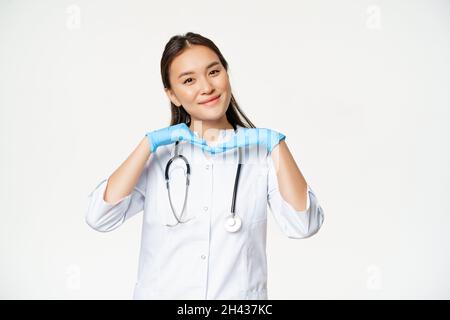 Image of smiling asian doctor, dermatologist shows healthy skin and posing cute at camera, standing in medical uniform over white background Stock Photo