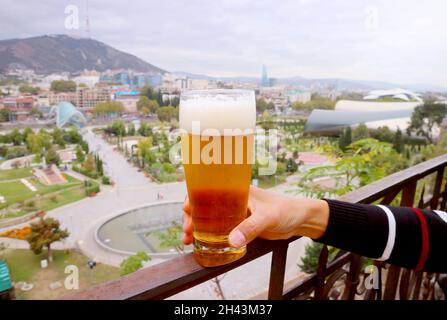 Hand Holding a Glass of Beer on the Balcony with Blurry City View in the Backdrop Stock Photo