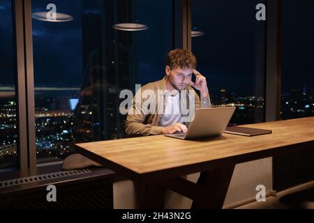 Thoughtful young man working online on computer late in office. Stock Photo