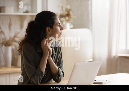 Pensive woman sit in kitchen with laptop looks into distance Stock Photo