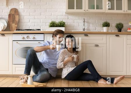Young couple holds cups sit on wooden floor in kitchen Stock Photo