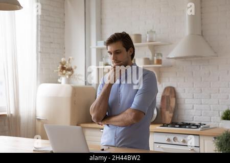 Man standing in kitchen staring at laptop thinks over task Stock Photo