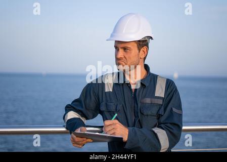 Professional young male port inspector in hardhat and protective uniform filling out papers while controlling shipment on pier Stock Photo