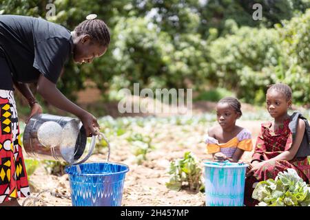 A tough young black African girl fills a water bucket with water from a metal watering can while her little sisters wait in silence for her to finish Stock Photo