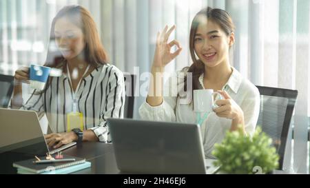 Charming business workers, businesswoman showing ok hand sign while holding coffee cup and having the meeting in meeting room. Stock Photo