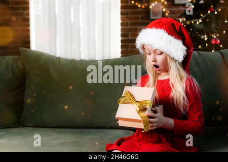 Small little shocked girl in red dress holds Christmas or New Year gift box present. Caucasian child surprised and say wow emotionally, exited because Stock Photo