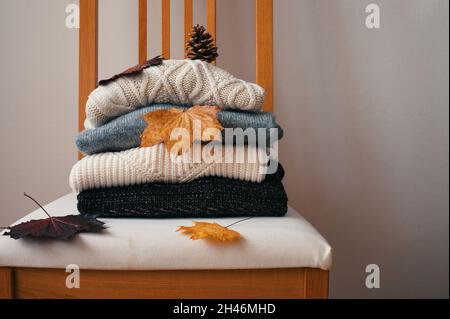 Stack of knitted warm sweaters on wooden chair with dry autumn leaves and pinecone Stock Photo