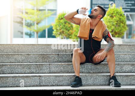 Fit handsome young man sitting on steps and drinking water after training outdoors Stock Photo