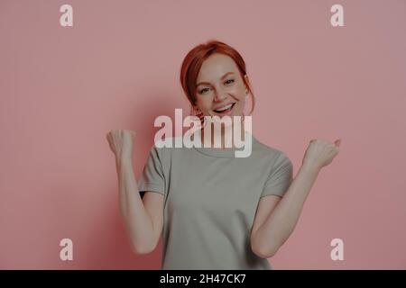 Overjoyed red-haired woman celebrating victory, doing winner gesture on pink background Stock Photo